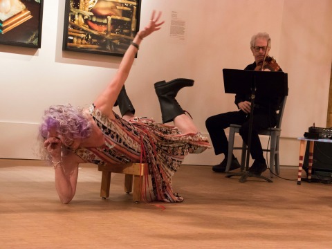 Celeste Miller poses lying crosswise on a stool with violinist behind her