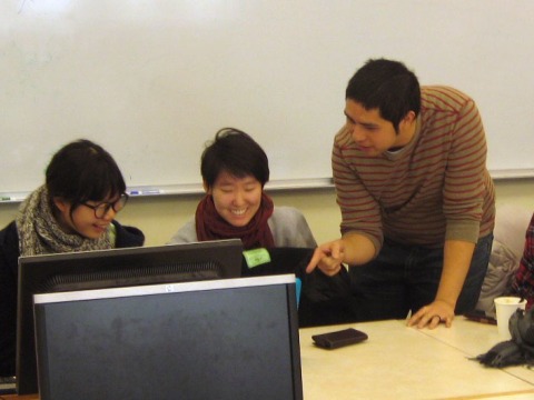 An instructor points to student work on a computer screen while two students look on.