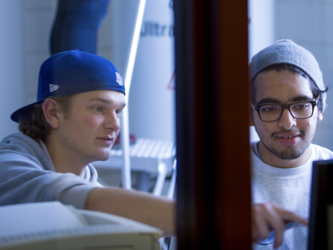 Two students look at a computer monitor and examine readings from the NMR machine in the background.