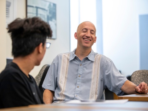 Jeff Blanchard, in a blue button-down shirt, laughs while sitting at a table with a student.