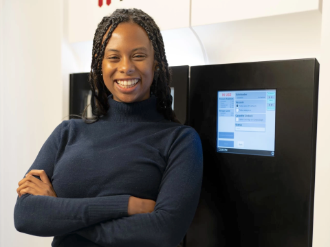 A young woman in a dark blue turtleneck and braided hair crosses her arms across her chest and smiles wildly. She stands in front of a large, cryogenic electron microscope.