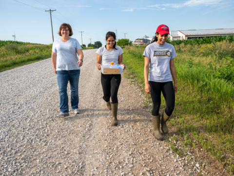 A woman and two students, also young women, walk toward the camera on a gravel road next to a field. They are conversing and smiling.