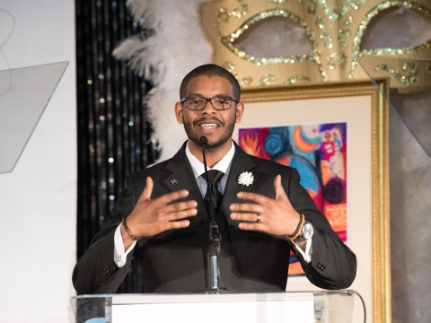 A man in a suit stands at a podium, gesturing with his hands as he delivers a speech. Behind him is a colorful abstract painting.