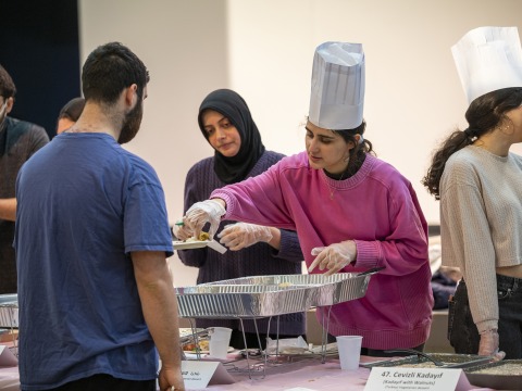 A young woman in a tall white chef's hat serves food to people waiting in line.