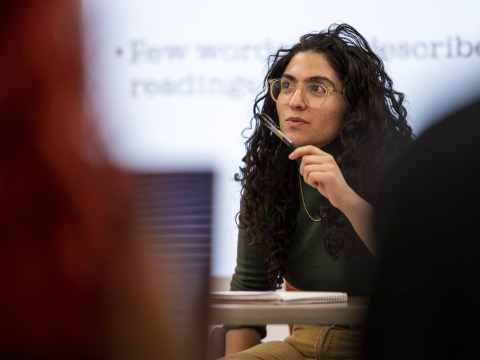 A woman in a green turtleneck and glasses looks intently off camera. In the foreground are the backs of students.
