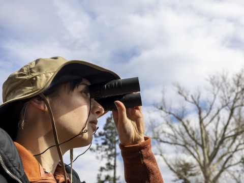 A woman in a beige, wide-brim hat holds a pair of binoculars to her eyes and gazes off camera. Behind her, leafless trees are backed by a blue, cloudy sky.