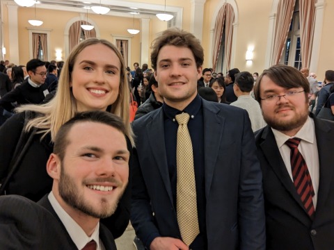 Four students in business attire with other conference attendees in the background.