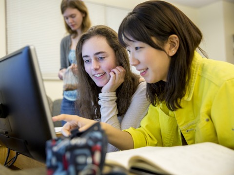 A girl with in a yellow jacket and a girl in a beige shirt laugh while pointing to something at the computer screen they share.