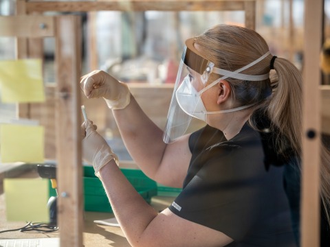 A woman in full protective gear holds up a small plastic saliva collection tube. She gestures with the tube as she speaks through a plexiglass window.