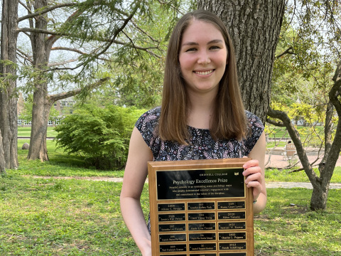 A young woman in a dress stands outdoors and smiles while holding a large plaque engraved with many names.