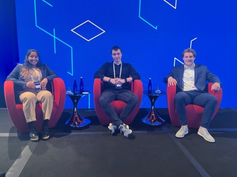 Three students with their conference badges, each in a red lounge chair in front of a graphic blue wall. Pictured are Mira Manchanda, Filip Matic, and Clayton Burton. 