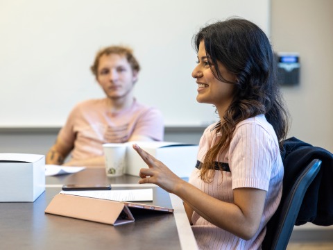 A student with black hair and pink shirt gestures with her hands as a student in the foreground looks intently in her direction.