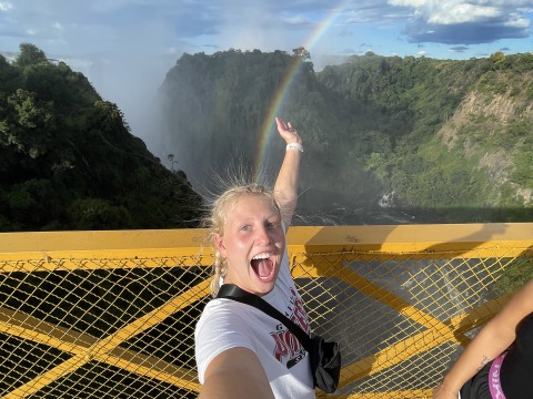 A young woman smiles while standing on a bridge over a deep gorge with a rainbow overhead
