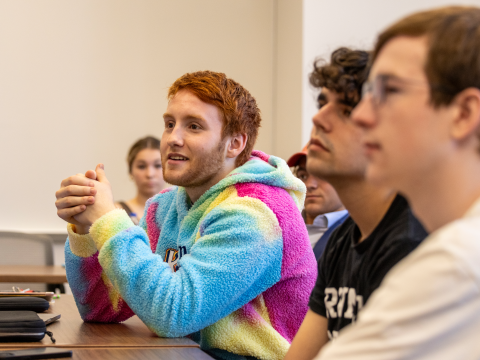 A ginger haired student with a colorful pink, blue, green sweater, hands clasped, looks off screen with a slight smile.