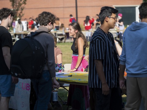 A focus on a student wearing pink velvet jeans. They talk to a student at a table outdoors.