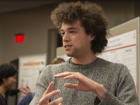Mikey, a young man with curly brown hair, gestures while giving a presentation at a poster session.