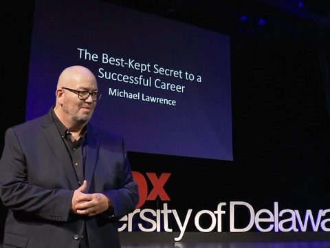A man in a suit clasps his hands together, presenting in front of a projector screen that reads, "The Best-Kept Secret to a Successful Career."