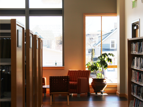 An open library space with open windows. On the left, an aisle of bookshelves and on the right, a shelf full of books.