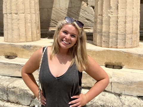 A young woman in sunglasses poses in front of ancient ruins in Greece.