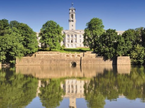 A white academic building with a single tower column. It is surrounded by trees and is behind a body of water.