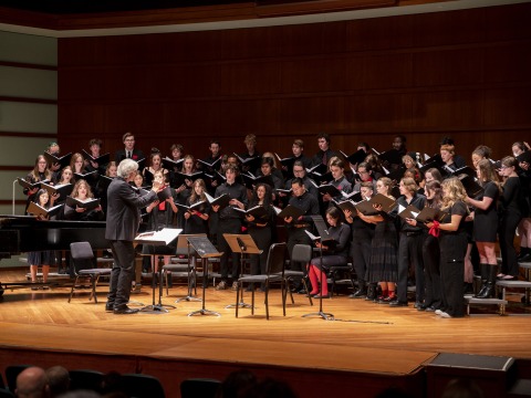 A choir sings on risers onstage, led by a male conductor