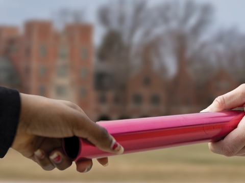 Two hands, one black and one white, hold opposite ends of a red baton. 