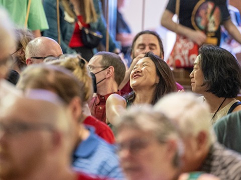alumni entering and sitting in an audience, central in the image are two people conversing, one laughing with her head thrown back