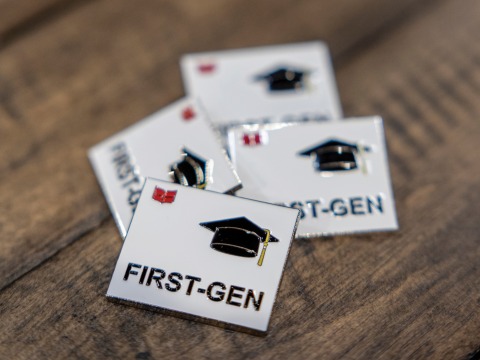 Four square white enamel pins sit on wooden planks The pins each have the word First-Gen and a mortar board in black with Grinnell College's red laurel leaves in upper left corner
