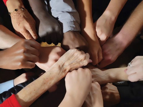 Hands in a wide variety of colors, clorhing, and decoration stack over each other in a circle.