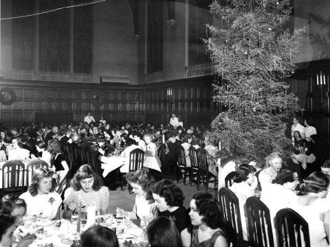 Black and white vintage photo showing women in long dresses enjoying a dinner in Main Hall under a huge Christmas tree