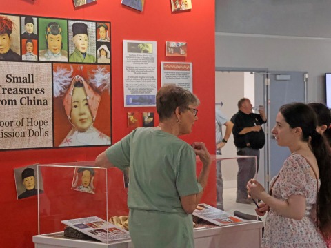 Three people stand at a glass museum case in front of a red wall with a large poster that reads 'Small Treasures from China: Door of Hope Mission Dolls'