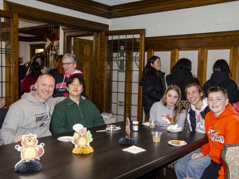 Five people sitting at a table and smiling. More people stand in the background chatting