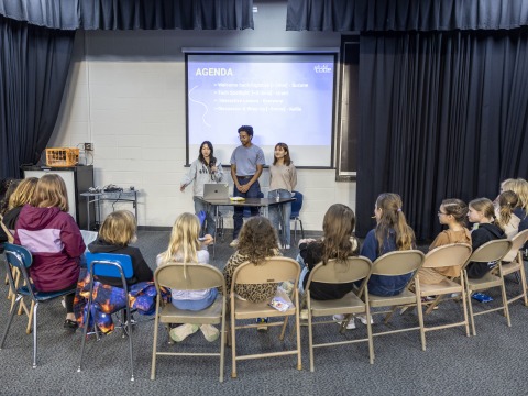 Three college students stand facing a semicircle of 15 middle school girls sitting in folding chairs