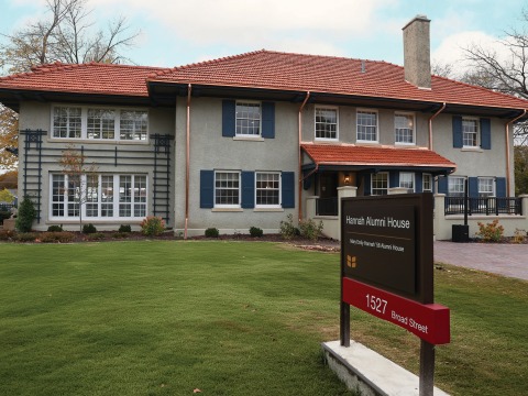 A two story stucco house with a red tile roof has a sign proclaiming it Hannah Alumni House 1527 Broad St
