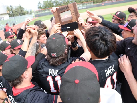 Baseball players wearing black uniforms with red accents celebrate in a group holding a wooden plaque