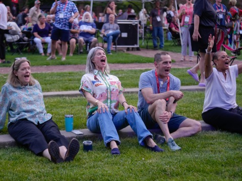 Four alums sitting on the grass cheering for the on stage performance.