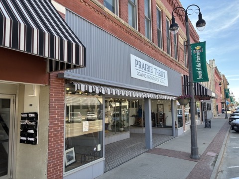 A row of shops with a white sign on a gray storefront reading "Prairie Thrift: Rewind. Rediscover. Reuse."