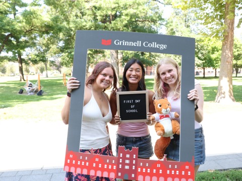 Three students pose for a photo holding a large grey photo frame and a sign that says First Day of School.