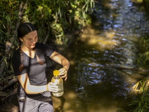 Student Millie Leonard stands in a small creek, holding a large, clear bottle containing a water sample.