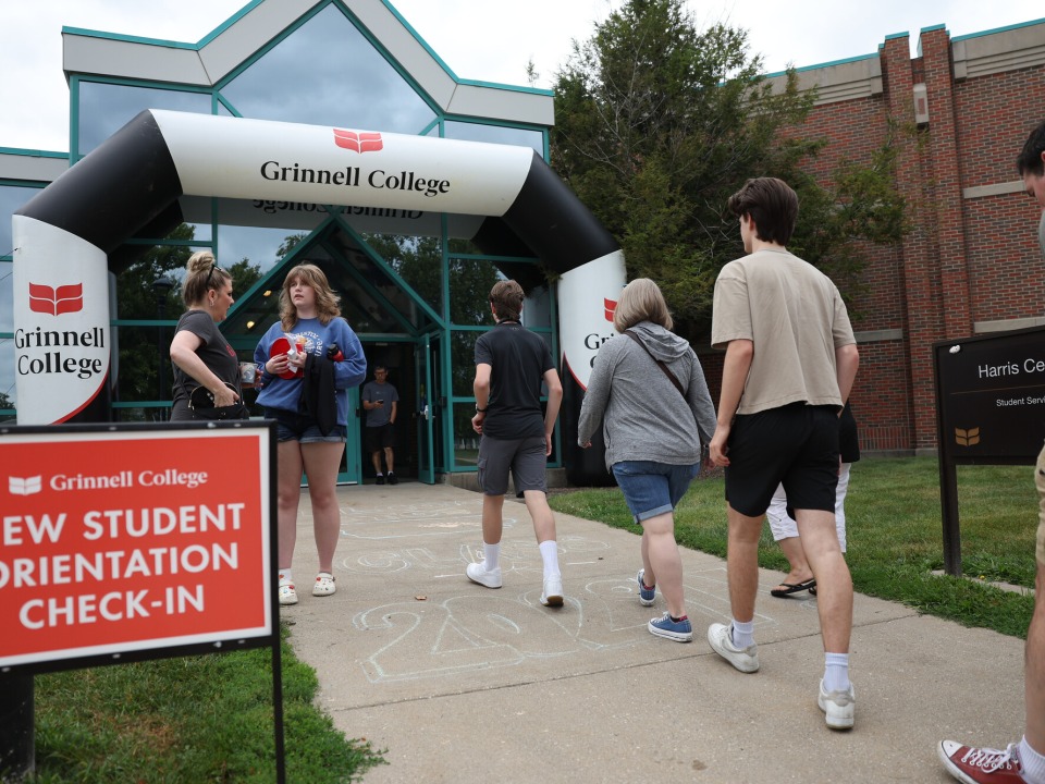People walk into a building through a black and white inflatable arch that is branded with "Grinnell College". A red sign with white text reads "New Student Orientation Check-In"