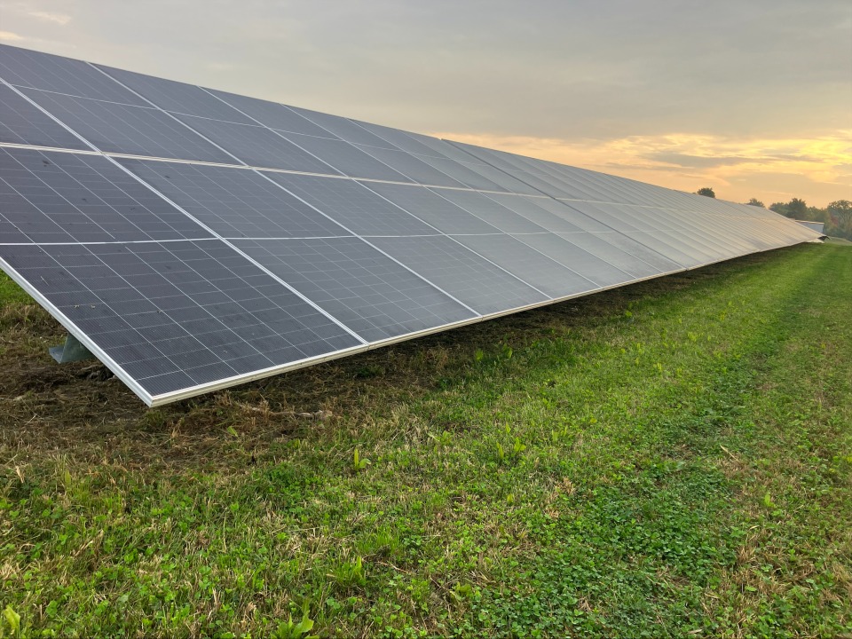 A bank of solar panels sits on a field with green grass with a cloudy sky above.