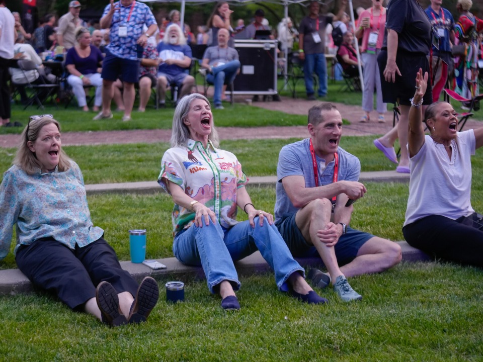 Four alums sitting on the grass cheering for the on stage performance.