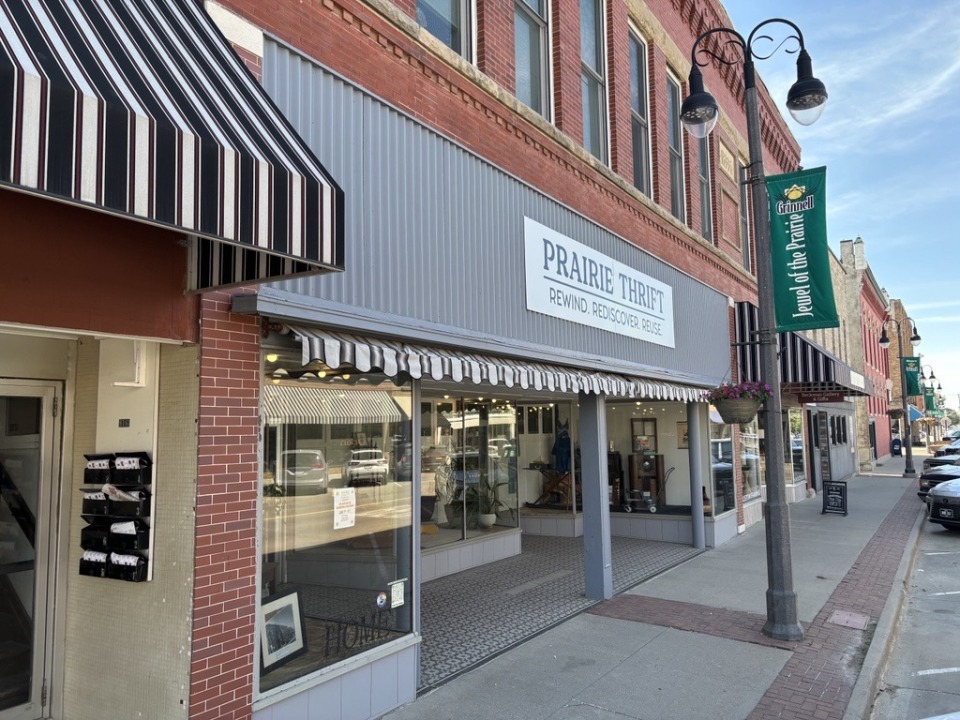 A row of shops with a white sign on a gray storefront reading "Prairie Thrift: Rewind. Rediscover. Reuse."
