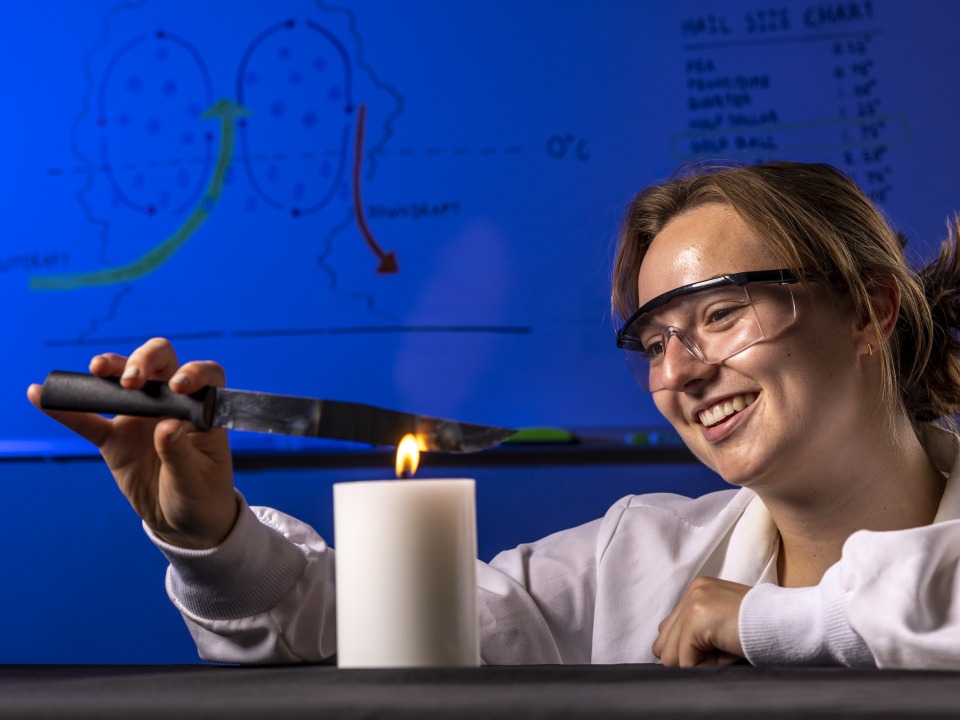 Anika Jane Beamer smiles as she performs a lab experiment by holding a small kitchen knife over a lit candle. She wears a white lab coat and protective glasses