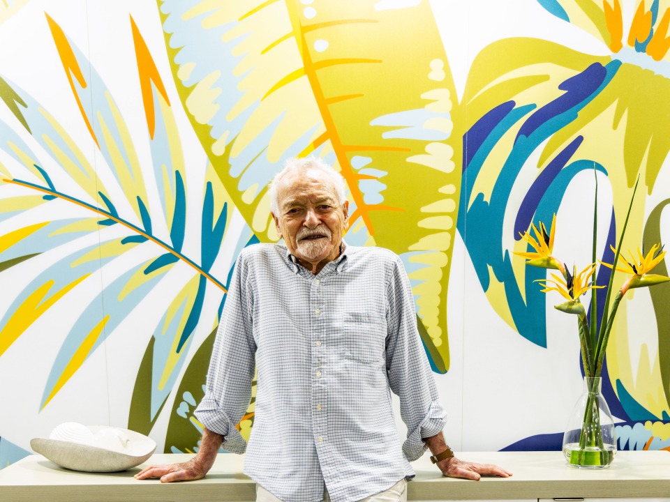 Tony Dalsimer, a senior person, wearing a striped button-up shirt leans on a white surface with a colorful tropical leaf mural and a vase with bird of paradise flowers in the background. 