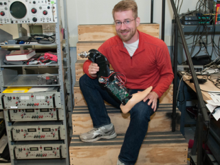  Brian Lawson holds a prosthetic leg. He is seated at the bottom of stairs in a room crowded with equipment