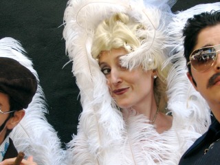 Three people: two blue-suited with mustaches and sunglasses, one with golden curls and large white feathers