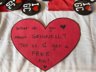 Red heart has black text asking what do you heart about grinnell? tell us and get a free pin. A group of GC pins appear beyond the heart