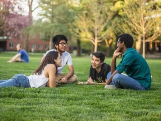 4 students face each other in a circle as they sit or lie on the grass