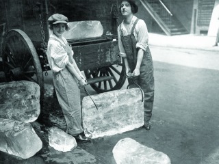 Two young women carry a large block of ice between them with large tongs